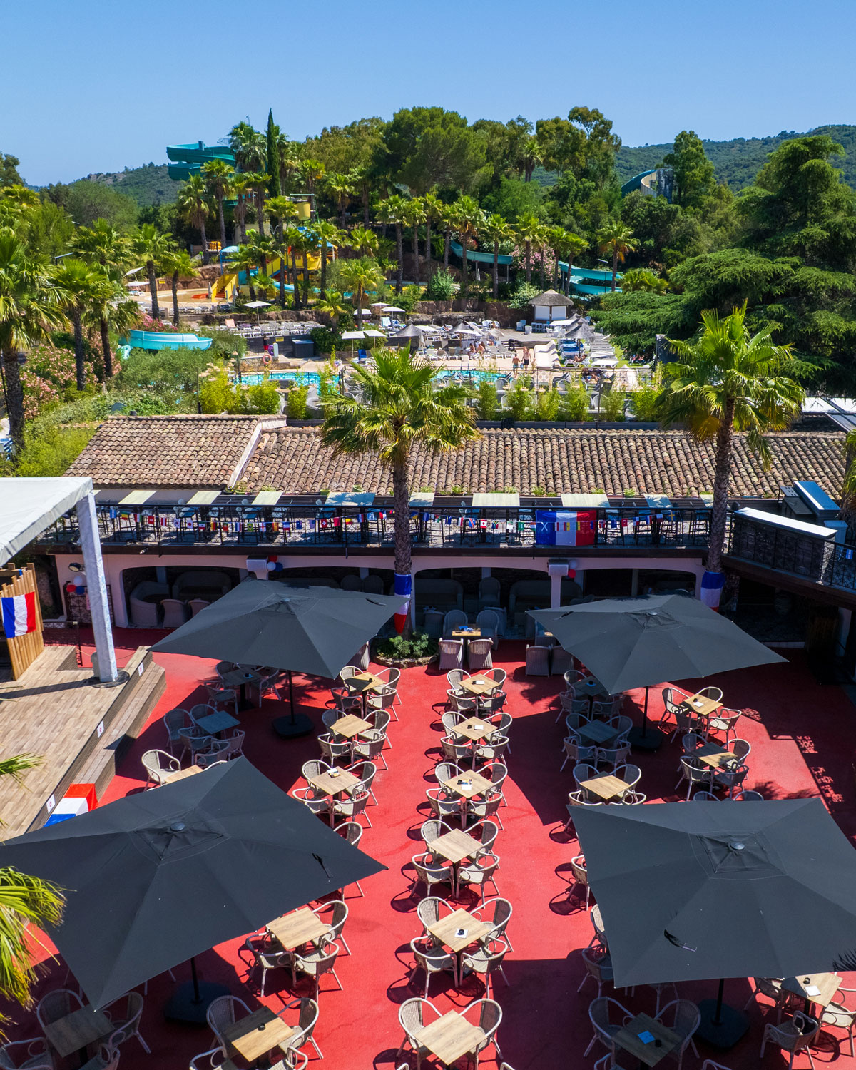 Terrasse der Bar von Esterel Caravaning unter Sonnenschirmen, im Hintergrund Palmen und die Rutschen der Wasserwelt