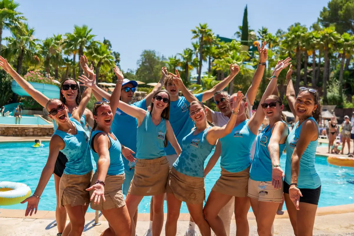 Das Team von Esterel Caravaning mit erhobenen Armen und strahlendem Lächeln am Pool unter Palmen